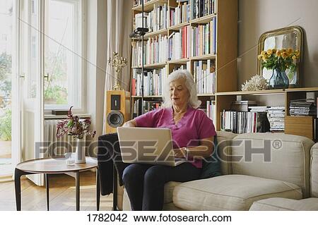 Stock Image - Senior woman using laptop on living room sofa. Fotosearch