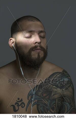 Serene bare chested man with beard and tattoos listening to music with earbuds View Large Photo Image Stock Photo - Serene bare chested man with beard and tattoos listening to music with earbuds. Fotosearch
