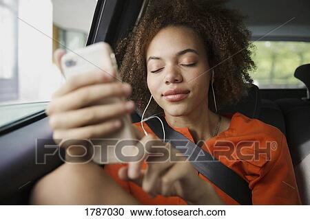 Stock Image - Serene young woman listening to music with headphones and mp3 player in back seat of car. Fotosearch
