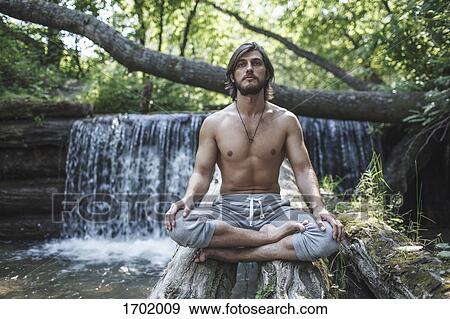 Stock Photo - Shirtless man sitting with crossed legs on logs against waterfall at forest. Fotosearch