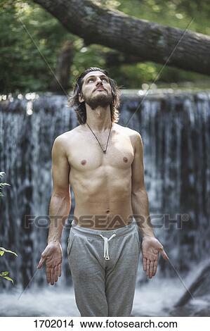 Shirtless young man looking up while standing against waterfall at forest View Large Photo Image Picture - Shirtless young man looking up while standing against waterfall at forest. Fotosearch