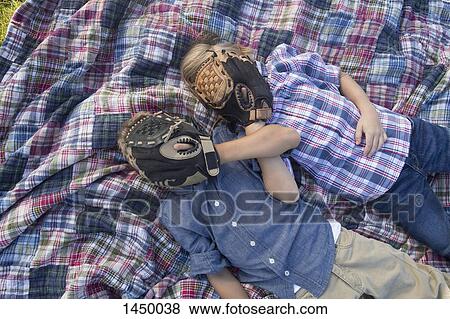 Stock Photo - Siblings covering face with baseball gloves while resting on blanket. Fotosearch
