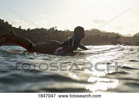 Stock Photo - Silhouette female surfer paddling out on sunny, sunset ocean. Fotosearch