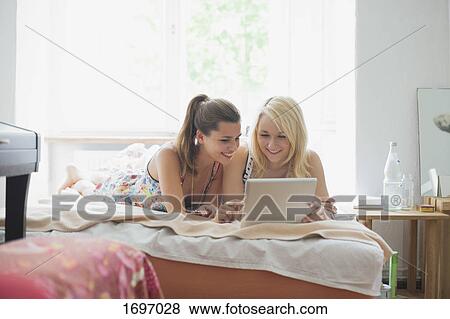 Smiling beautiful young female friends using digital tablet while lying on bed at home View Large Photo Image Stock Photo - Smiling beautiful young female friends using digital tablet while lying on bed at home. Fotosearch