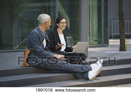 Smiling business colleagues with laptop discussing while sitting on steps in city View Large Photo Image Stock Photo - Smiling business colleagues with laptop discussing while sitting on steps in city. Fotosearch