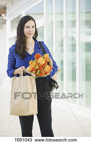 Smiling confident woman holding flowers bouquet and paper bag while standing in city View Large Photo Image Stock Image - Smiling confident woman holding flowers bouquet and paper bag while standing in city. Fotosearch