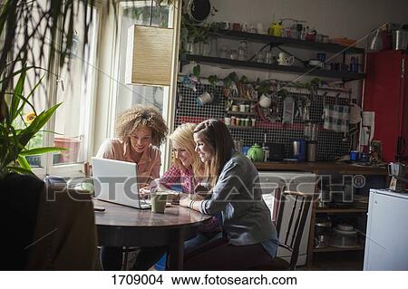 Smiling female friends using laptop at table by window View Large Photo Image Picture - Smiling female friends using laptop at table by window. Fotosearch