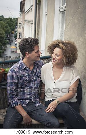 Smiling friends talking while sitting by wall in balcony View Large Photo Image Stock Photo - Smiling friends talking while sitting by wall in balcony. Fotosearch