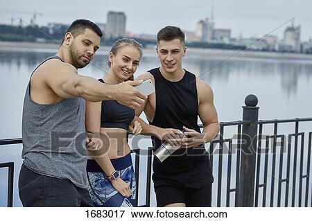Stock Photo - Smiling male athlete taking selfie with friends while standing against lake. Fotosearch