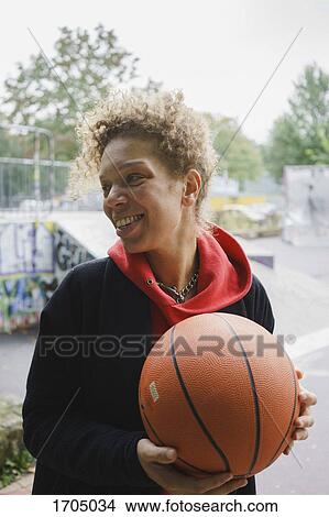 Picture - Smiling mid adult woman holding basketball while standing at park. Fotosearch