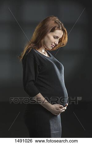 Stock Photograph - Smiling pregnant woman holding stomach against gray background. Fotosearch
