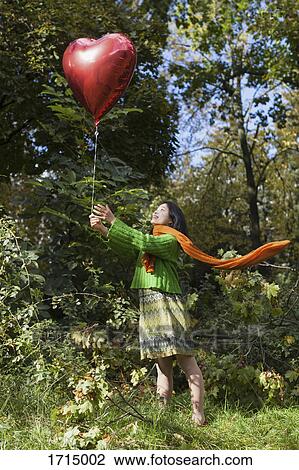 Smiling woman holding red heart shape balloon while standing on field against trees at park View Large Photo Image Stock Image - Smiling woman holding red heart shape balloon while standing on field against trees at park. Fotosearch
