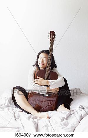 Smiling woman with closed eyes embracing guitar while sitting on bed at home View Large Photo Image Stock Photo - Smiling woman with closed eyes embracing guitar while sitting on bed at home. Fotosearch