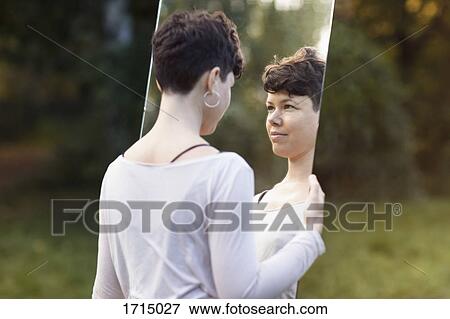 Stock Photo - Smiling woman with short hair looking in mirror while standing at park. Fotosearch