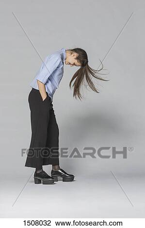 Stock Image - Smiling woman with tousled hair and hands in pockets standing against gray background. Fotosearch
