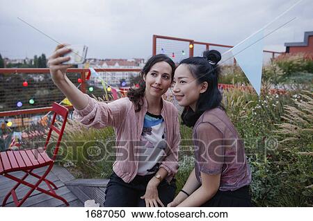 Stock Image - Smiling women taking selfie with smart phone on patio. Fotosearch