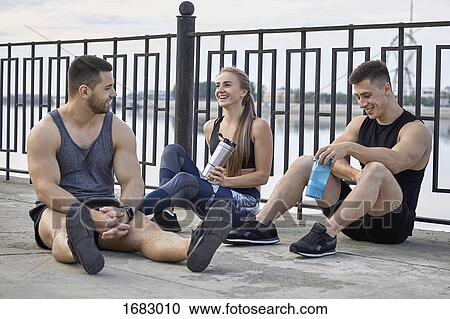Stock Image - Smiling young athletes taking break while sitting on footpath by railing. Fotosearch