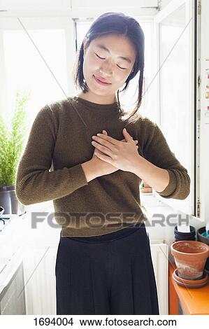 Picture - Smiling young woman with hands clasped on heart standing against window. Fotosearch
