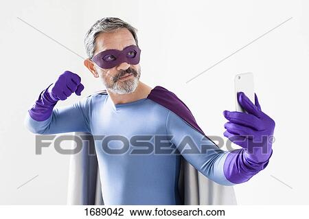 Stock Image - Superhero taking selfie from smart phone while standing against white background. Fotosearch
