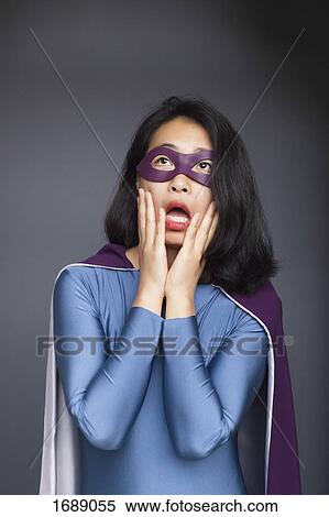 Stock Photography - Surprised woman dressed in superhero costume standing with head in hands against gray background. Fotosearch