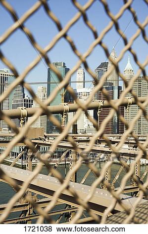 Stock Photo - Tall buildings seen through chainlink fence on Brooklyn Bridge, New York City, New York, USA. Fotosearch