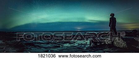 Stock Photograph - Teenage boy watching Northern Lights from rock, Reykjanesbaer, Iceland. Fotosearch