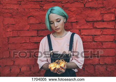 Stock Photography - Teenage girl holding illuminated string lights while standing against brick wall. Fotosearch
