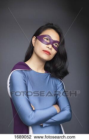 Stock Image - Thoughtful female superhero standing with arms crossed against gray background. Fotosearch