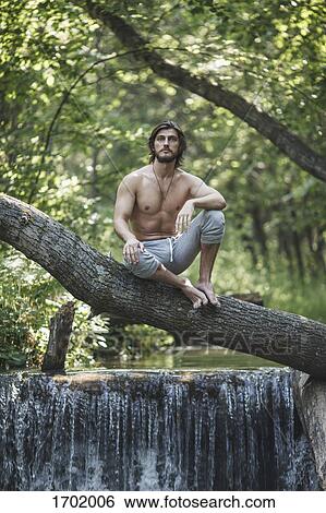 Stock Photograph - Thoughtful shirtless man sitting on branch over waterfall at forest. Fotosearch