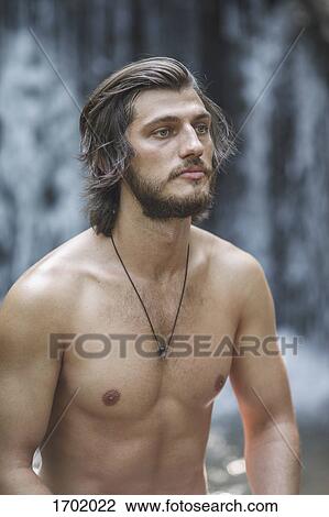 Stock Image - Thoughtful shirtless young man looking away while standing against waterfall at forest. Fotosearch