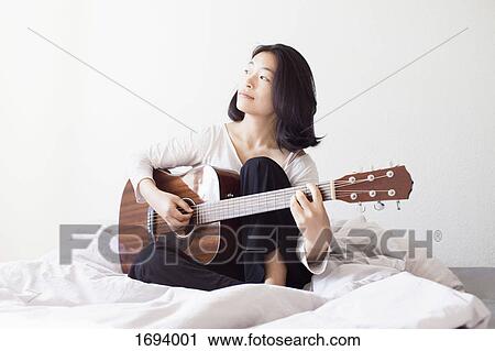 Stock Image - Thoughtful woman playing guitar while sitting on bed at home. Fotosearch
