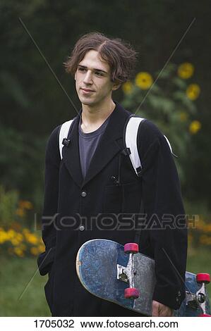 Stock Image - Thoughtful young man carrying skateboard while standing at park. Fotosearch