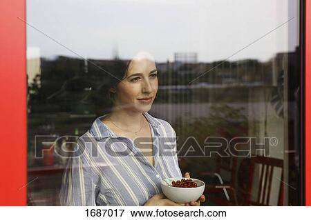 Stock Photo - Thoughtful young woman holding bowl with red currants while looking through window. Fotosearch