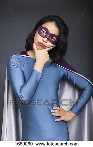 Stock Image - Thoughtful young woman wearing superhero costume standing with hand on hip against gray background. Fotosearch