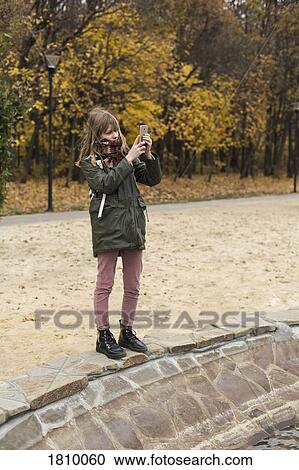 Stock Image - Tween girl using camera phone in autumn park. Fotosearch