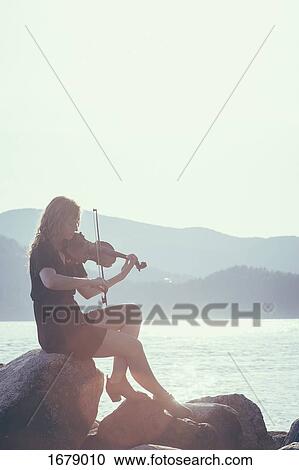 Stock Image - Woman playing violin while sitting on rock at lakeshore against clear sky. Fotosearch