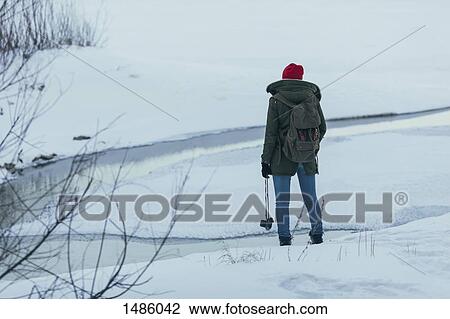 Stock Image - Woman with backpack holding camera while standing on field during winter. Fotosearch