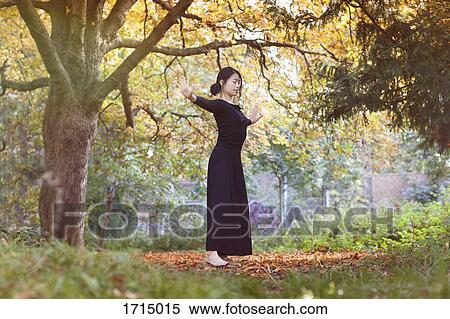 Stock Photography - Woman with closed eyes and arms outstretched standing on field against trees at park. Fotosearch