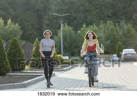 Stock Photo - Women friends riding bicycle and push scooter on road. Fotosearch