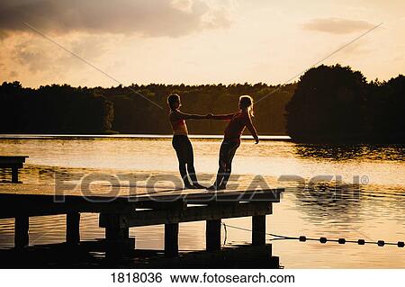 Stock Photograph - Women practicing acro yoga on tranquil sunset lake dock. Fotosearch