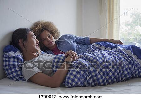 Stock Image - Young couple lying on bed against white wall by window at home. Fotosearch