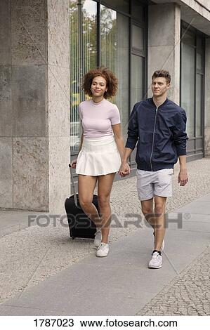 Stock Image - Young couple with suitcase walking on urban sidewalk. Fotosearch