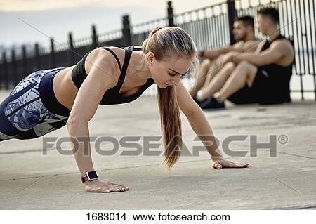 Young female athlete practicing push-ups against male friends sitting on footpath View Large Photo Image Picture - Young female athlete practicing push-ups against male friends sitting on footpath. Fotosearch