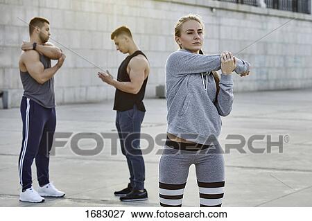 Young female athlete stretching with friends standing in background on footpath View Large Photo Image Stock Photo - Young female athlete stretching with friends standing in background on footpath. Fotosearch