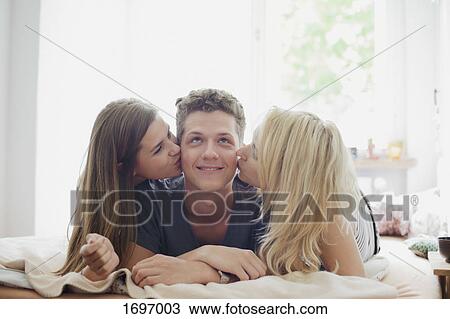 Young female friends kissing handsome man lying on bed at home View Large Photo Image Stock Image - Young female friends kissing handsome man lying on bed at home. Fotosearch