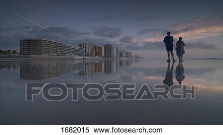 Stock Photography - Young man and woman standing on shore at beach against sky during sunset, Daytona, Florida, USA. Fotosearch