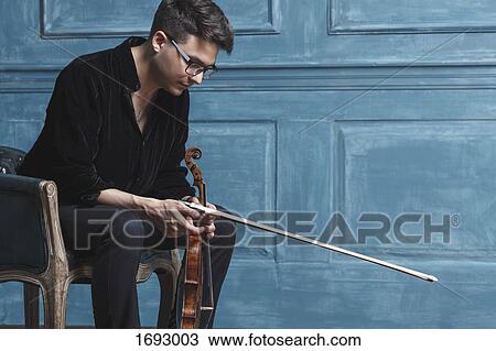 Young man holding violin while sitting on chair by blue wall View Large Photo Image Stock Image - Young man holding violin while sitting on chair by blue wall. Fotosearch