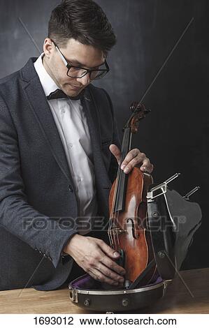 Stock Image - Young man keeping violin in case at table against black background. Fotosearch