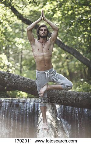 Stock Image - Young man standing in tree pose on log against waterfall at forest. Fotosearch