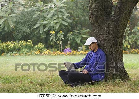 Young man using laptop while sitting by tree on grassy field at park View Large Photo Image Stock Image - Young man using laptop while sitting by tree on grassy field at park. Fotosearch